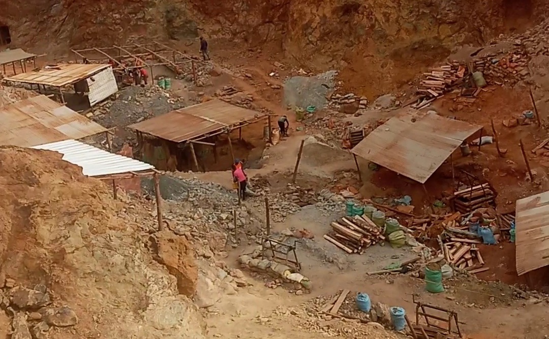 person writing in a notebook beside by an iPad, laptop, printed photos, spectacles, and a cup of coffee on a saucer