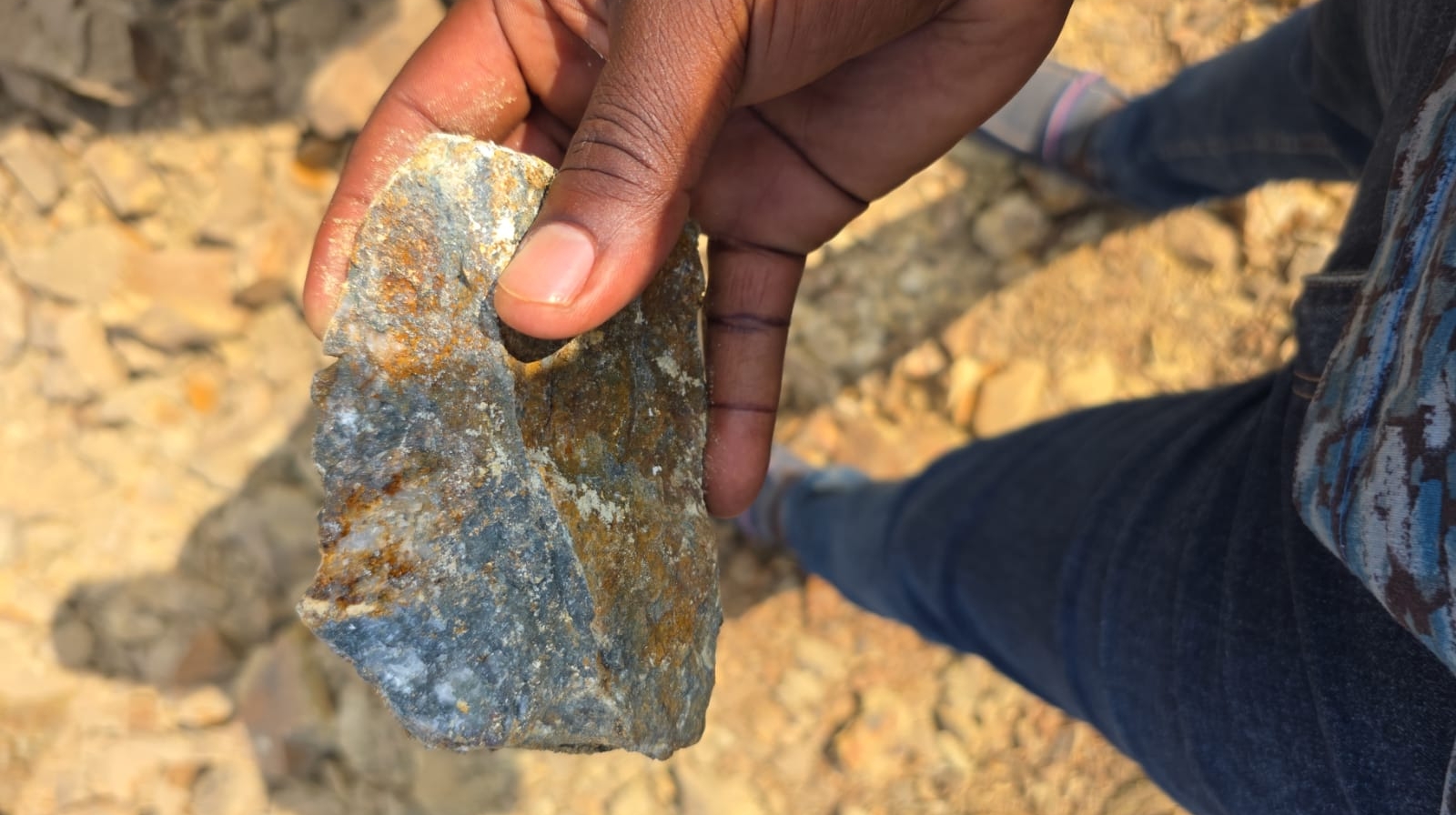 person writing in a notebook beside by an iPad, laptop, printed photos, spectacles, and a cup of coffee on a saucer